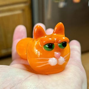 以下が含まれることがあります： 緑色の目をしたオレンジ色のガラス製の猫の置物。猫は手に乗っています。白いひげがあります。