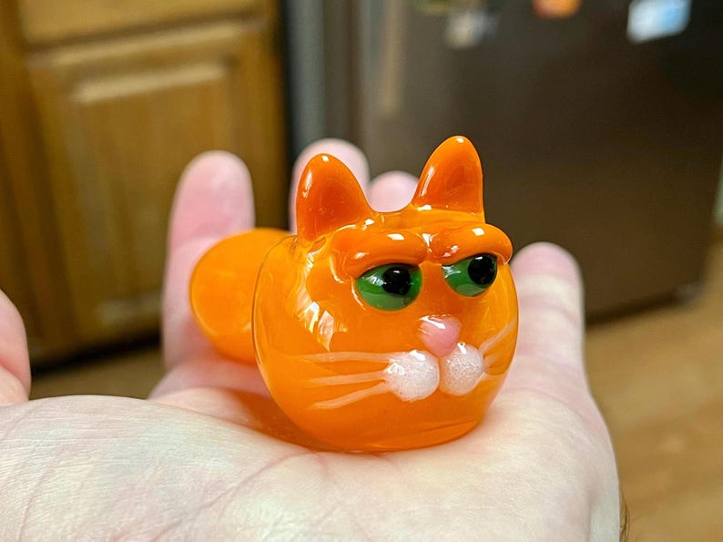以下が含まれることがあります： 緑色の目をしたオレンジ色のガラス製の猫の置物。猫は手に乗っています。白いひげがあります。