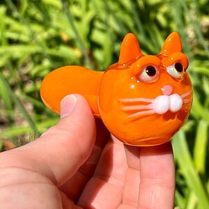 以下が含まれることがあります： 白い目とひげのあるオレンジ色のガラス製の猫のパイプ。パイプは猫の頭と体の形をしています。