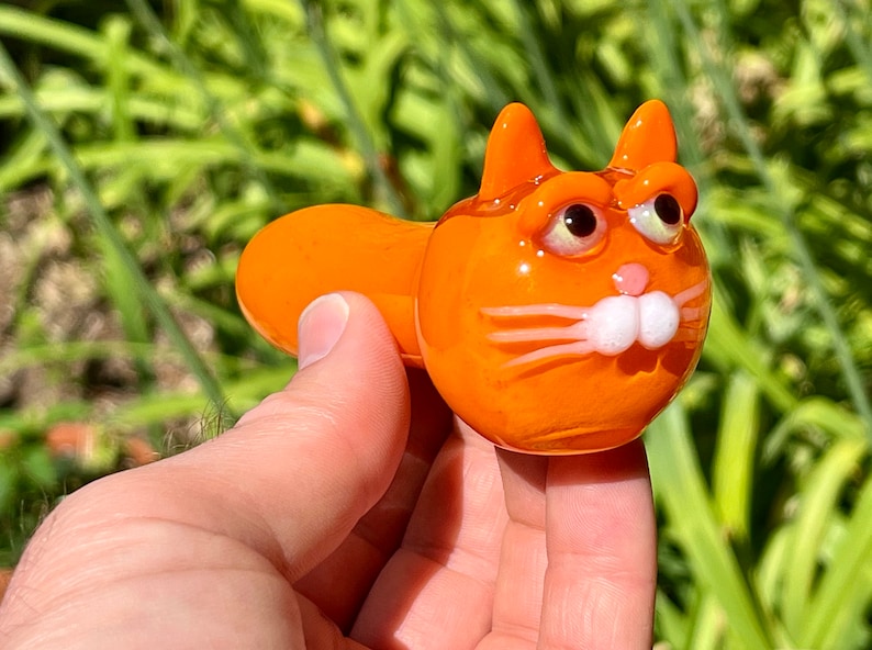 以下が含まれることがあります： 白い目とひげのあるオレンジ色のガラス製の猫のパイプ。パイプは猫の頭と体の形をしています。