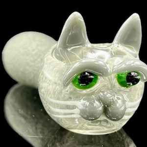May include: A white glass cat pipe with green eyes and white whiskers. The pipe has a textured surface and a round shape.