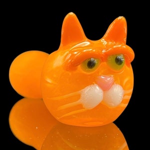 以下が含まれることがあります： 白いひげと大きな緑色の目をしたオレンジ色のガラス製の猫のパイプ