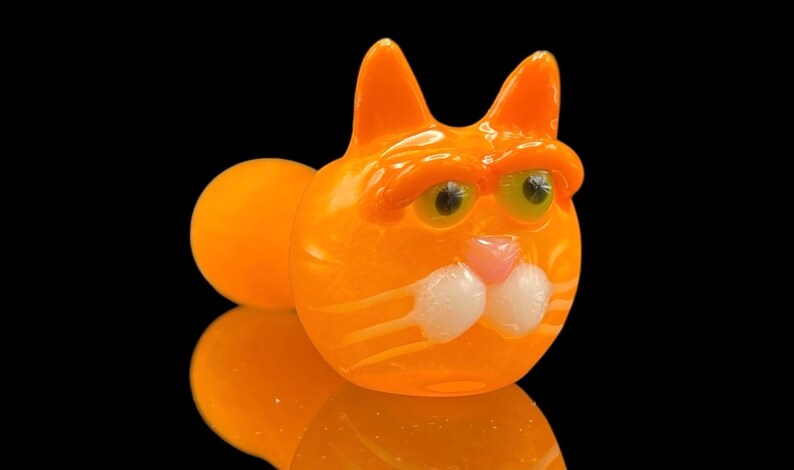 以下が含まれることがあります： 白いひげと大きな緑色の目をしたオレンジ色のガラス製の猫のパイプ