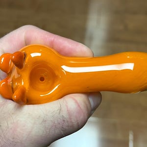 以下が含まれることがあります： 丸いボウルと広がったマウスピースを持つオレンジ色のガラスパイプ。パイプはボウルに3つの小さな突起があるユニークなデザインです。