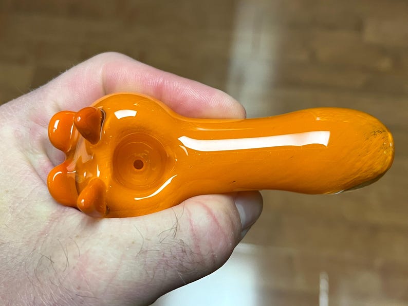 以下が含まれることがあります： 丸いボウルと広がったマウスピースを持つオレンジ色のガラスパイプ。パイプはボウルに3つの小さな突起があるユニークなデザインです。