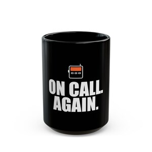 May include: A black ceramic mug featuring a white graphic of a pager with an orange screen. Below the pager icon, the text ON CALL AGAIN. is printed in bold white capital letters. The mug has a cylindrical shape with a smooth, glossy finish.