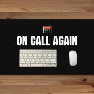May include: A black rectangular desk mat featuring a white graphic of a pager above the white text ON CALL AGAIN. A white wireless keyboard and a white computer mouse are positioned on the mat surface, creating a workspace setup.