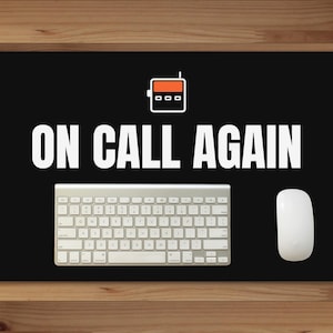 May include: A black rectangular desk mat featuring a white graphic of a pager with an orange screen and the white text ON CALL AGAIN. A silver wireless keyboard and a white computer mouse are placed on top of the mat.