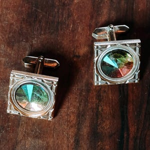 Può includere: Un paio di gemelli argentati con una cornice quadrata e un gioiello ovale multicolore. I gemelli hanno un perno rettangolare e sono posizionati su uno sfondo di venature del legno scuro. I gioielli riflettono sfumature di verde, arancione e blu.