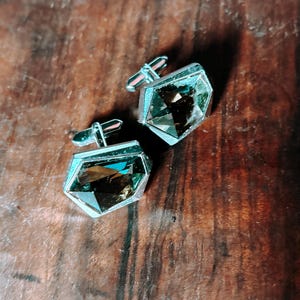 Può includere: Un paio di gemelli argentati con pietre di cristallo fumé geometriche e sfaccettate. I gemelli sono appoggiati su una superficie di legno scuro, evidenziando le loro qualità riflettenti e il loro design elegante.