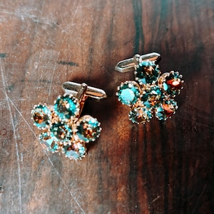 Può includere: Un paio di gemelli a forma di fiore con steli in metallo color oro. Ogni gemello presenta un gruppo di pietre preziose color verde acqua, marrone e trasparenti. I gemelli sono appoggiati su una superficie di legno scuro.
