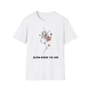 May include: White t-shirt featuring a black and white illustration of a woman's profile adorned with roses and butterflies. The text "BLOOM WHERE YOU ARE" is printed in black below the illustration. A casual, everyday wear tee.