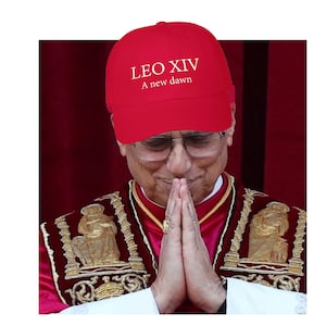 May include: A red baseball cap with the text "LEO XIV A new dawn" in white. The cap is worn by a person wearing glasses and a red and gold robe, with hands clasped in prayer.