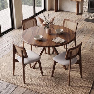 Può includere: Un tavolo da pranzo rotondo in legno con venature marrone scuro, accompagnato da quattro sedie con sedute imbottite grigio chiaro e schienali in paglia di Vienna intrecciata. Un vaso in ceramica con fiori secchi e due ciotole sul piano.