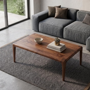 May include: A rectangular wooden coffee table with tapered legs, featuring a smooth grain surface. On the table sits a small beige bowl, a stack of three books, and a small potted succulent in a metallic gold container, all resting on a rug.