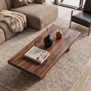 May include: A rectangular coffee table made of dark brown wood with a visible grain pattern. On the table sits a black ceramic vase with bare branches, a glass reed diffuser with wooden sticks, and a book titled Stelogeture of Luxury.