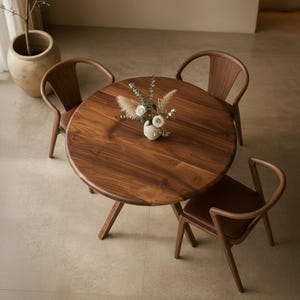 Può includere: Un tavolo da pranzo rotondo in legno con tre sedie abbinate. Il tavolo ha una finitura marrone scuro e una base a tre gambe. Le sedie hanno schienali curvi e sedili in pelle marrone. Un piccolo vaso di fiori è sul tavolo.
