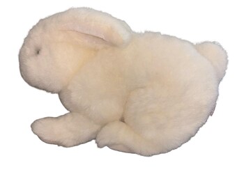 May include: A plush stuffed animal in the shape of a rabbit. The rabbit is covered in soft, off-white, cream-colored fur. It is depicted in a resting, seated position with its ears folded back against its head and a small, rounded tail visible.