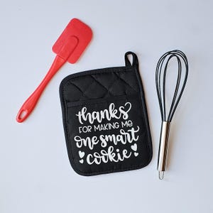 May include: Black quilted oven mitt with the text "Thanks? For making me one smart cookie" in white lettering. The mitt is accompanied by a red silicone spatula and a silver whisk.