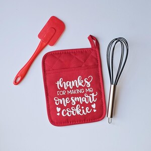 May include: Red quilted hot pad with the text "thanks for making me one smart cookie" in white with two small hearts on either side of the word "cookie". A red silicone spatula and a silver whisk are also pictured.