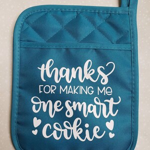 May include: Teal quilted oven mitt with a white design that says "Thanks for making me one smart cookie" with two white hearts on either side of the word "cookie".