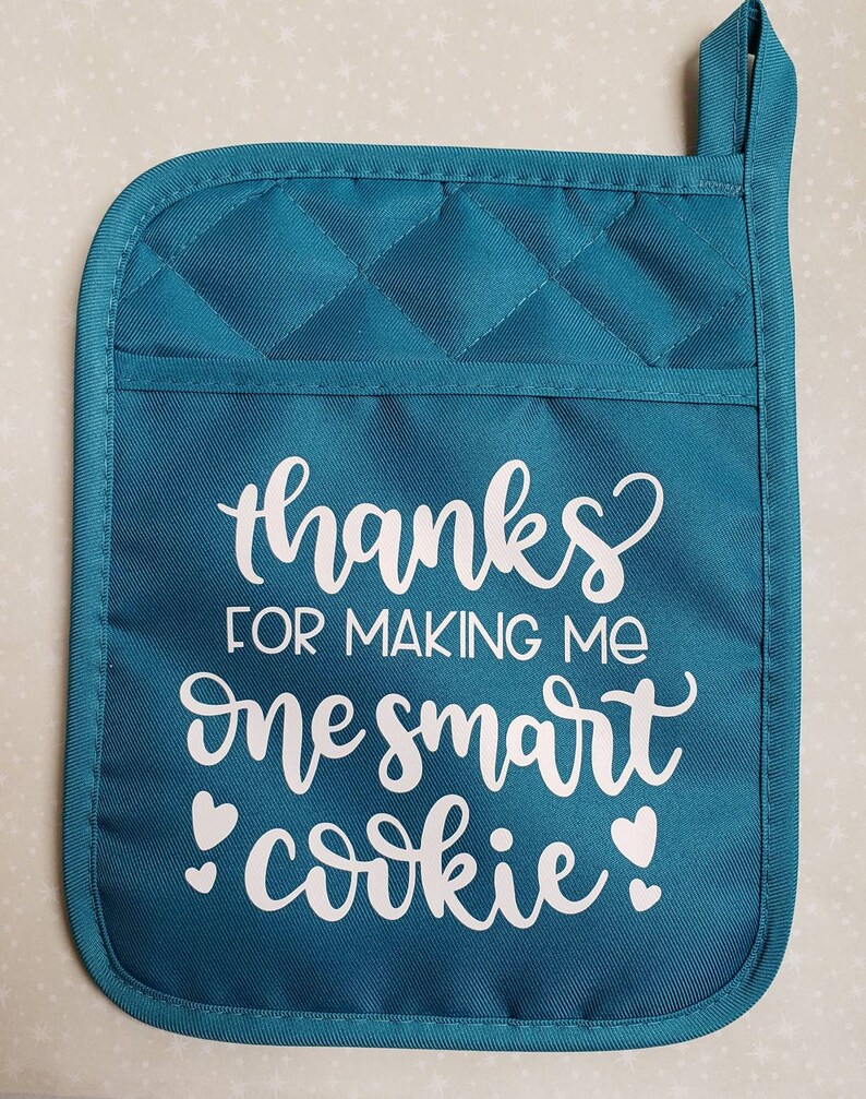 May include: Teal quilted oven mitt with a white design that says "Thanks for making me one smart cookie" with two white hearts on either side of the word "cookie".