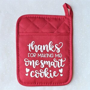 May include: Red quilted oven mitt with a white design that says "Thanks for making me one smart cookie" with two small hearts on either side of the word "cookie".