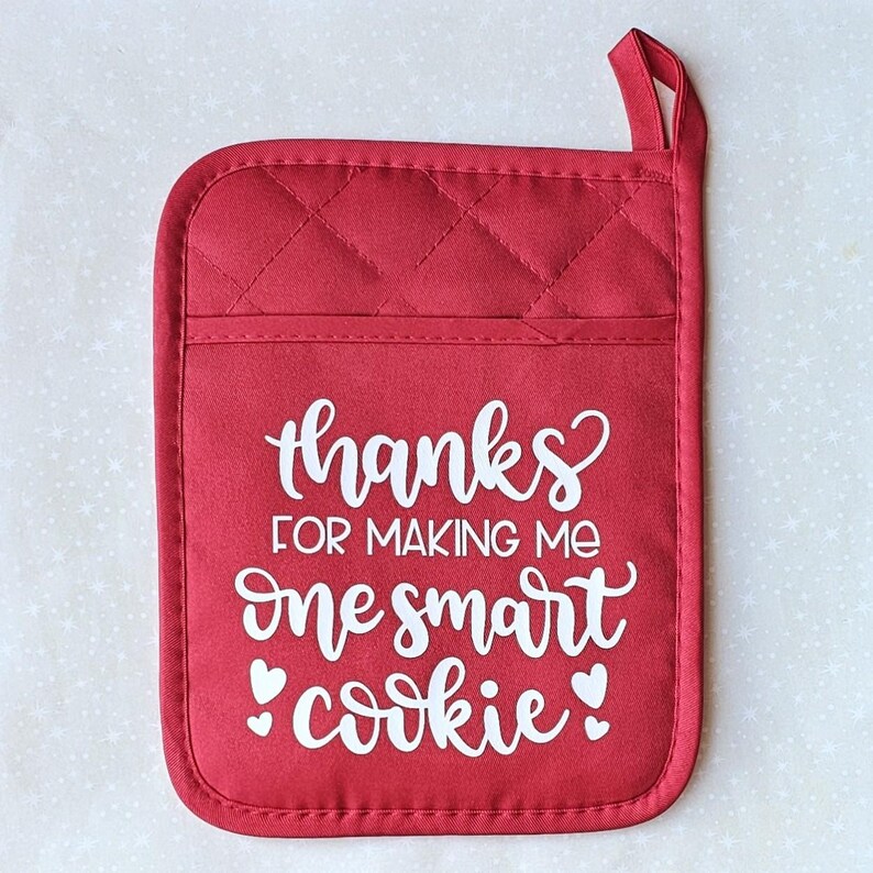 May include: Red quilted oven mitt with a white design that says "Thanks for making me one smart cookie" with two small hearts on either side of the word "cookie".