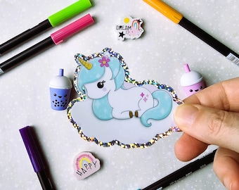 May include: A sparkly iridescent sticker featuring a cartoon unicorn with a blue mane and tail, lying on a cloud. The sticker is surrounded by colorful markers, erasers, and other small items.