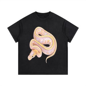 Könnte beinhalten: Schwarzes T-Shirt mit einer Aquarellillustration einer Schlange. Die Schlange ist in Rosa-, Gelb- und Weißtönen gehalten und detailliert dargestellt. Das T-Shirt hat einen klassischen Rundhalsausschnitt und kurze Ärmel.