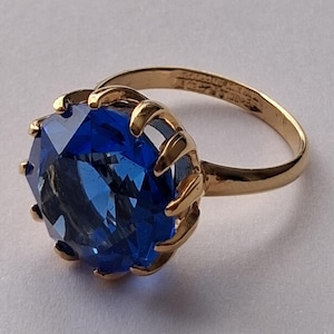 May include: A gold ring featuring a large, round, faceted blue gemstone held in place by a multi-prong setting. The band is smooth and polished gold, with visible hallmarks stamped on the inner surface of the ring band.