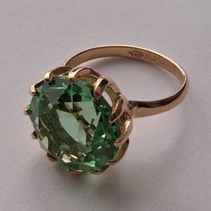 Peut inclure: Une bague en or ornée d'une grande pierre précieuse ronde et facettée de couleur vert clair, maintenue par un sertissage à griffes. L'anneau est lisse et poli, avec un poinçon visible gravé sur la surface intérieure du métal.