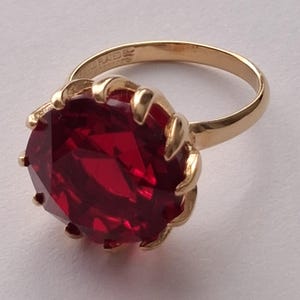 Könnte beinhalten: Ein vergoldeter Ring mit einem großen, runden, facettierten tiefroten Edelstein in einer Krappenfassung. Die Innenseite der Ringschiene ist mit dem Text GOLD PLATED graviert und zeigt eine polierte metallische Oberfläche sowie einen leuchtenden Stein.