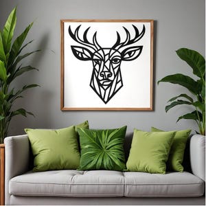 May include: A framed wall art piece featuring a geometric black deer head design on a white background. The artwork is encased in a light brown wooden frame. The image is displayed above a gray sofa with green throw pillows.
