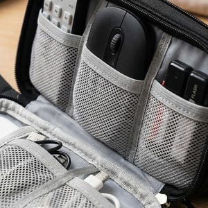 May include: Black zippered electronics organizer with multiple mesh pockets. Contains a computer mouse, remote control, flash drives, and charging cables. The case has a handle for easy carrying.