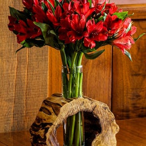 Può includere: Un vivace bouquet di fiori rossi con steli e foglie verdi in un vaso di vetro trasparente. Il vaso è posto su una base di legno intagliato con una forma naturale e organica. Lo sfondo è una parete e un tavolo di legno.