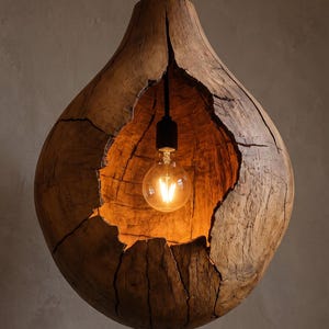 Puede incluir: Una lámpara colgante en forma de gota hecha de una pieza de madera marrón natural ahuecada con grietas visibles y una textura rugosa. Una bombilla de cristal transparente cuelga de un cable negro dentro de la abertura central de la madera.