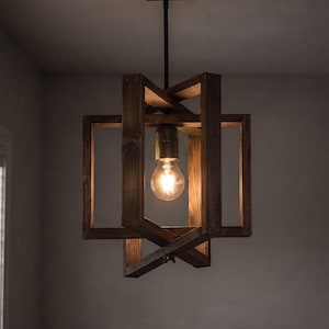 Puede incluir: Una lámpara colgante con un marco de madera marrón oscuro construido con vigas rectangulares que se cruzan. Un casquillo de tono latón sostiene una bombilla de cristal transparente, suspendida de un soporte de techo de madera a juego.