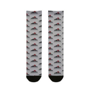 May include: A pair of grey socks featuring a repeating pattern of a mountain scene and a red maple leaf. The text "MONASHEE" is printed above the mountain scene. The socks have black heels and toes.
