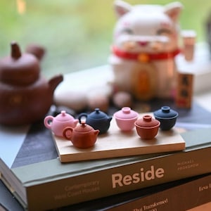 Könnte beinhalten: Eine Sammlung miniaturer Keramik-Teekannen und -Tassen in verschiedenen Farben, darunter Rosa, Braun und Marineblau, angeordnet auf einem Holztablett. Eine braune Teekanne und eine weiße Katzenfigur im Hintergrund. Das Tablett liegt auf einem Stapel Bücher.