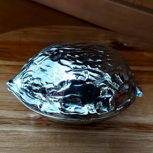 May include: A silver-coloured, walnut-shaped nutcracker. The nutcracker has a textured surface and is resting on a wooden surface. The nutcracker is a decorative item for the kitchen or dining room.