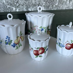 May include: A collection of four white ceramic kitchen storage jars with lids. Each jar has a scalloped edge and is adorned with colourful fruit and floral patterns, including apples, pears, and blueberries. The jars are of different sizes.