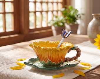 May include: A ceramic tea cup and saucer set shaped like a sunflower with yellow petals and a green leaf base. The cup features a textured brown branch-like handle and includes a silver-toned spoon topped with a small blue and white butterfly.