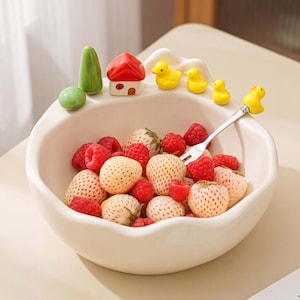 May include: A white ceramic bowl with an irregular rim, featuring decorative miniature figures of a green tree, a red-roofed house, and three yellow ducks on the edge. The bowl is filled with red raspberries and white strawberries, with a metal fork.