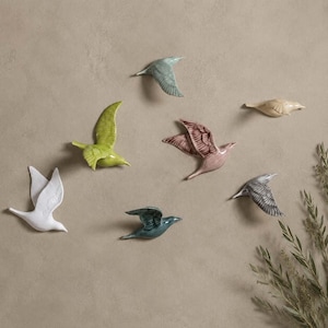 May include: A collection of seven ceramic bird wall sculptures in various colours including white, lime green, teal, dusty rose, light blue, and beige, each depicted in a different flight pose with detailed wing textures and smooth finishes.