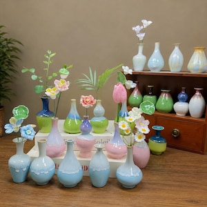 May include: A collection of small ceramic vases in various pastel and vibrant colors, including blue, pink, green, and white. The vases feature smooth, glazed finishes, some with drip-style patterns, and are displayed on white books and a wooden shelf.