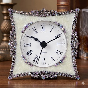 Peut inclure: Une horloge de table carrée avec un cadran blanc, des chiffres romains noirs et des aiguilles noires. Le mot QUARTZ est imprimé sur le cadran. Le cadre est de couleur crème avec un motif tourbillonnant et orné de nombreux petits cristaux violets.