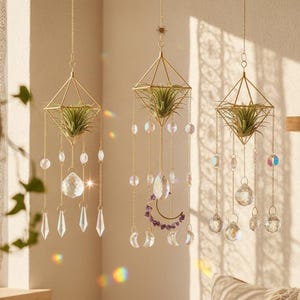 May include: Three hanging geometric gold-toned metal planters, each containing a green air plant. The planters are suspended by chains adorned with clear glass crystals, faceted prisms, and a crescent moon charm with purple stone accents.