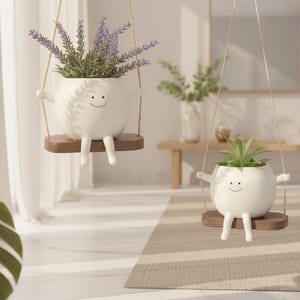 May include: Two white ceramic planters shaped like round figures with smiling faces, arms, and legs. Each planter sits on a small, rectangular dark brown wooden swing suspended by thin ropes, holding green plants, including purple lavender.