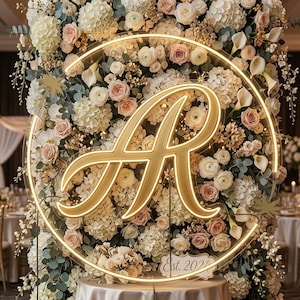 May include: A circular neon sign featuring a large, glowing gold letter A in a cursive script, mounted on a dense floral backdrop of white and blush pink roses, hydrangeas, and calla lilies, with a small plaque at the bottom reading Est. 2024.